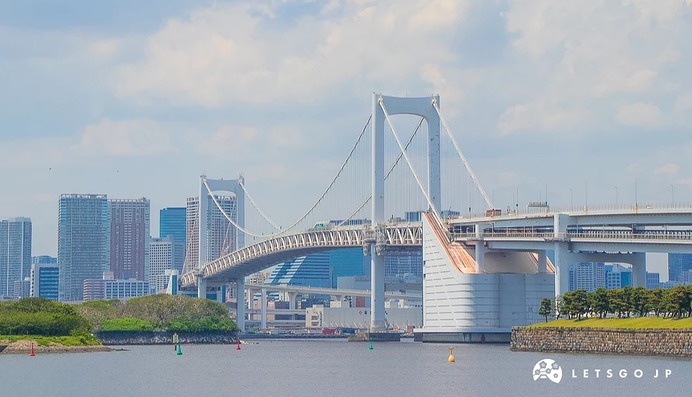 東京彩虹大橋全景 白色懸索彩虹大橋橫跨海面,背景為城市高樓與藍天白雲。