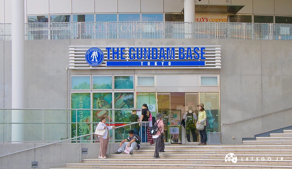 鋼彈基地東京入口 THE GUNDAM BASE 專賣店玻璃門入口,門前有幾位遊客正在階梯上歇息。