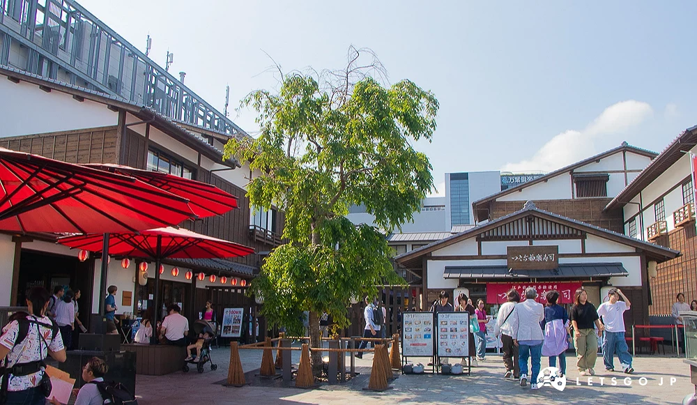 豐洲千客萬來廣場 日式傳統木造建築廣場,中央種植綠色樹木,左側設有大型紅色遮陽傘。