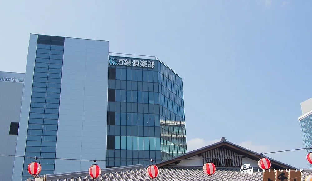 萬葉俱樂部大樓外觀 玻璃帷幕現代大樓頂端標示「万葉倶楽部」,前方為日式瓦片屋頂與紅皮燈籠。