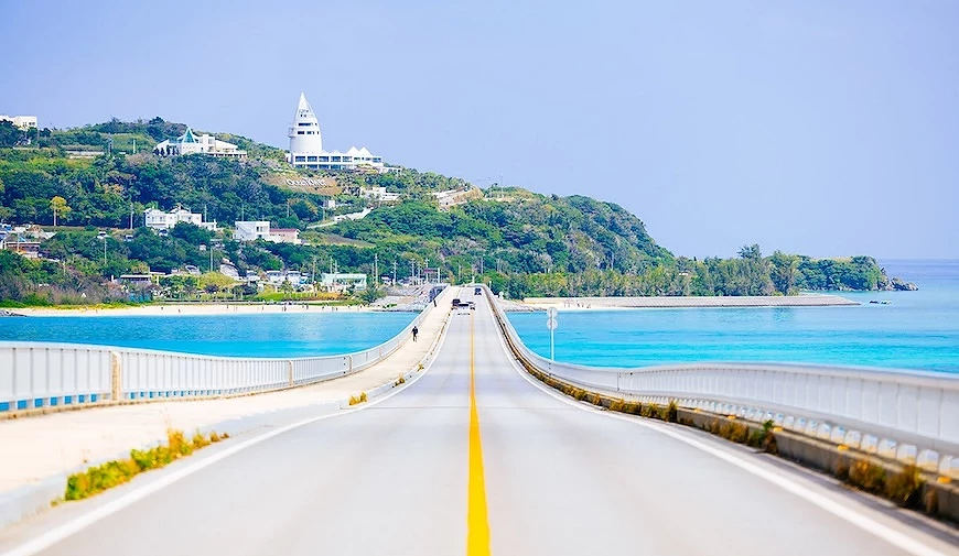 筆直跨海橋道路延伸至綠丘上的白色尖塔建築,兩側為藍綠色海域