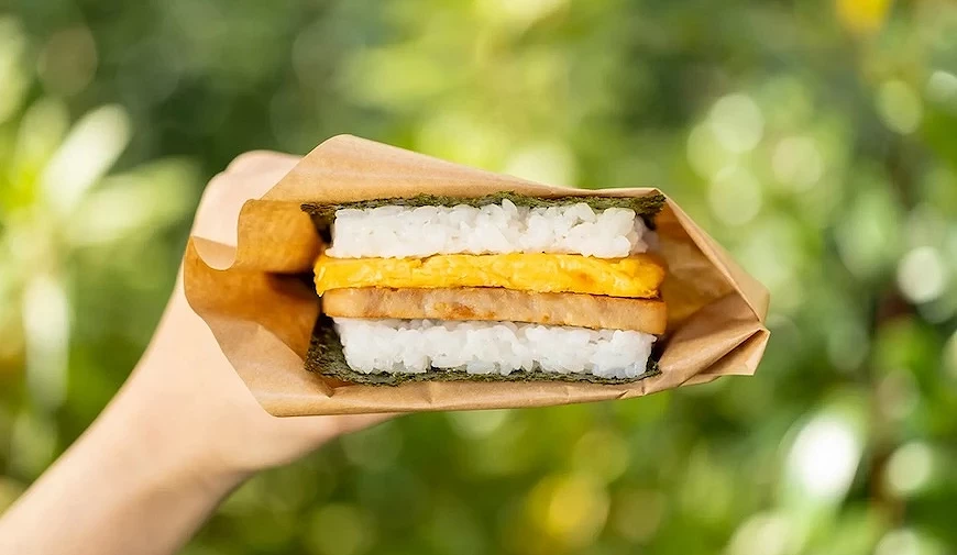 手持以海苔與白飯夾住煎蛋及豬排的飯糰三明治，以牛皮紙包覆，戶外綠意背景