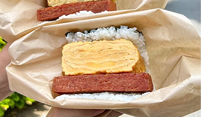 雙手各持一份以牛皮紙包覆的午餐肉玉子飯糰，戶外綠樹與道路為背景