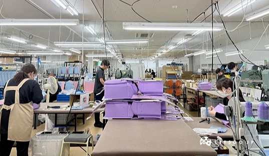 敞明亮的工廠內擺放多台縫紉機與工作桌,數名工作人員正在不同工位製作日本小學生書包。