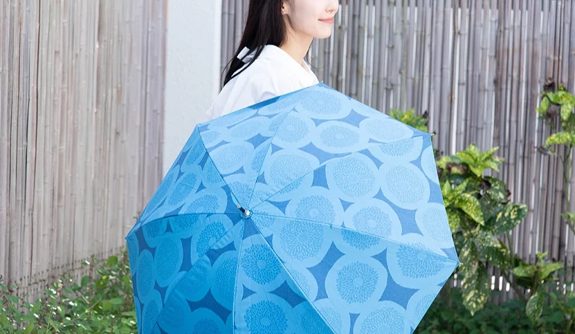 女子手持蓝色几何花纹阳伞站在户外庭院中,伞面采渐层蓝色设计,展现日式工艺感与优雅质感。