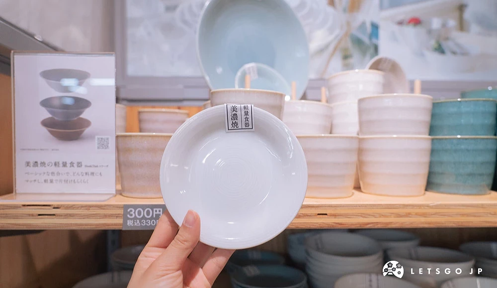 手持 Standard Products 美浓烧白色轻量餐盘,后方陈列多款不同颜色与尺寸的美浓烧餐具