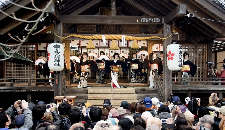 日式神社舞台表演,舞者穿着和服起舞,白色灯笼与绳饰点缀,游客聚集观赏祭典。