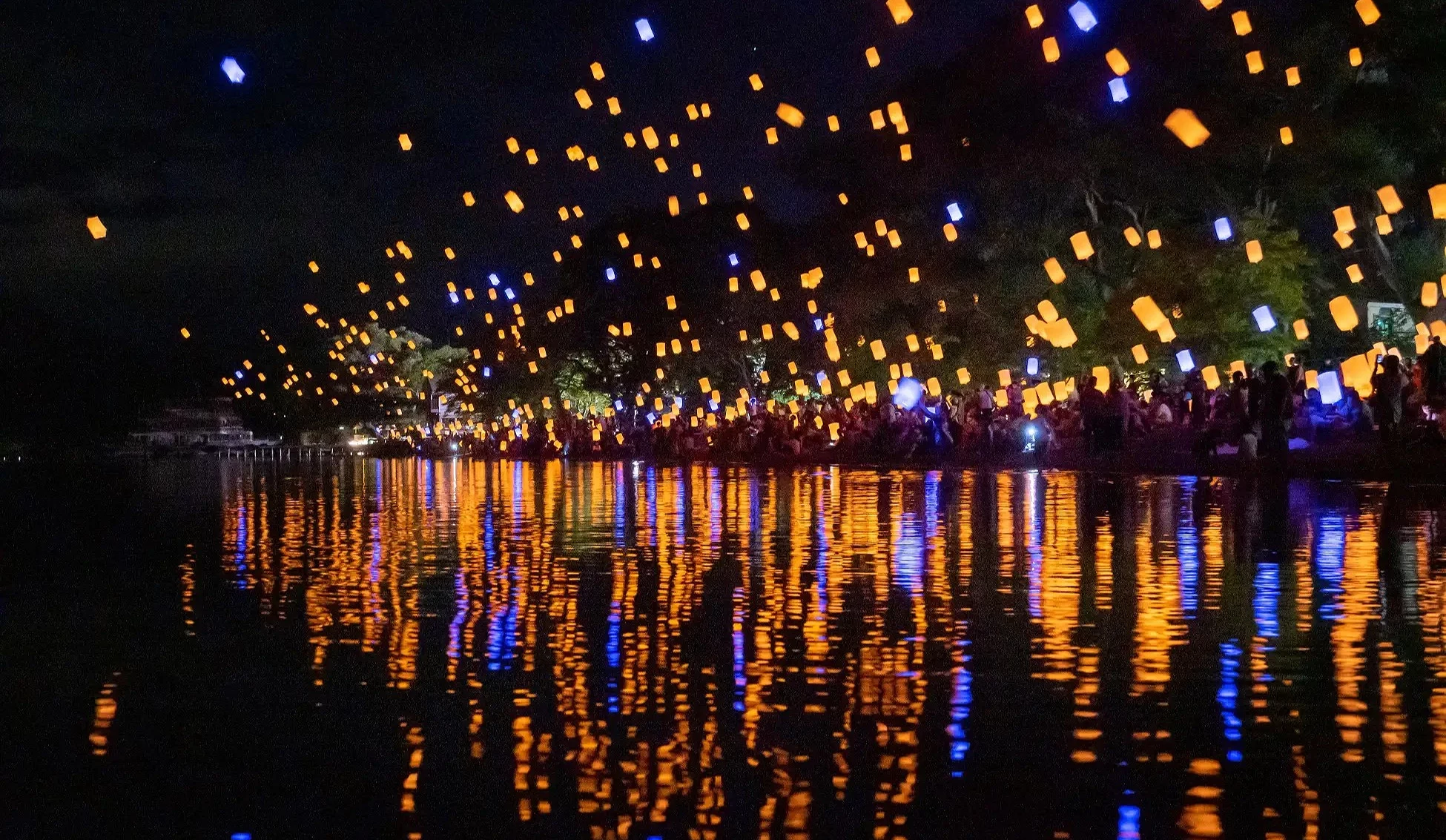 夜空中無數天燈升空,湖面倒映橘藍光影,人群在岸邊觀賞。
