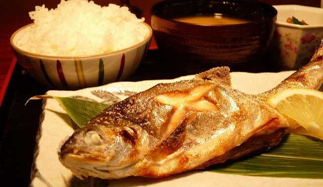 日式烤魚定食近景。前方是一整條表皮酥脆的烤魚,附一塊黃檸檬和一片竹葉。後方配有一大碗白飯、一碗味噌湯和一小碟配菜,擺放在黑色托盤上