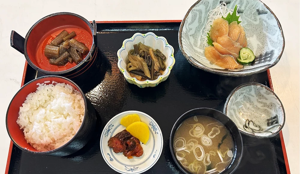 從上方俯瞰精緻的日式傳統定食套裝,擺放在帶紅邊的黑色矩形托盤上。菜色包含一大碗白飯、一碗味噌湯、一碟生魚片刺身、兩碗不同樣式的日式小菜,以及一小碟黃色和紫色的醃漬物