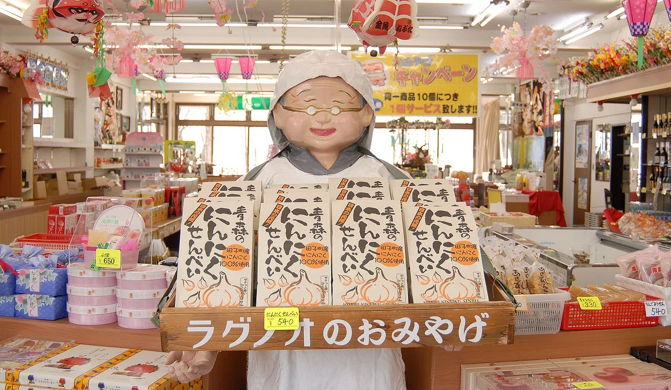 當地土產店的室內特寫,展示當地特產「ラグノオのおみやげ」(RAGUENEAU的伴手禮)。一個身穿白袍、戴帽子和眼鏡的人形玩偶站在印有產品標誌和圖案的商品盒子前方,盒子擺放在一個木箱裡。人形玩偶掛著微笑,周圍環繞著掛滿吊飾的商店背景。